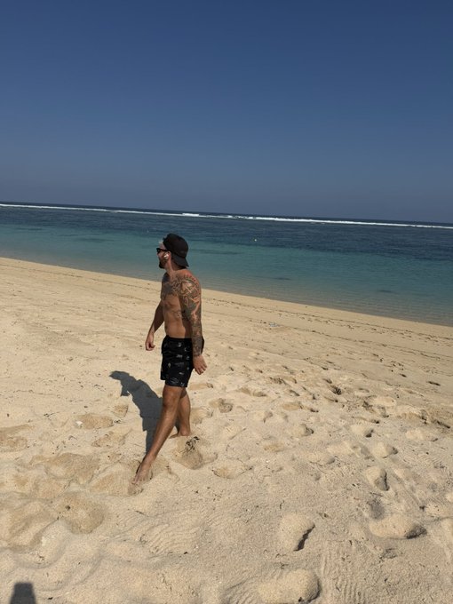 A shirtless man wearing black shorts and a backward cap walks along a sandy beach beside clear turquoise water.