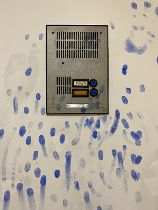 A gray intercom or doorbell unit mounted on a white wall covered with blue handprints and paint splatters.