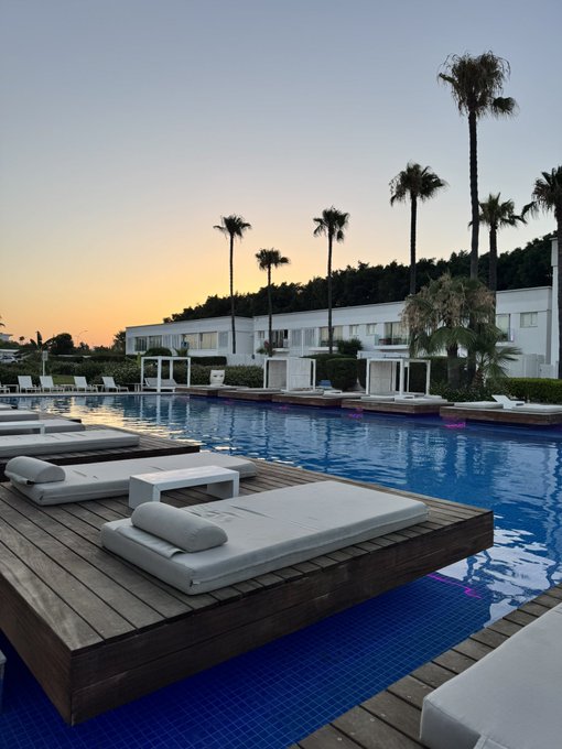 A serene resort pool at sunset with white lounge beds on wooden decking over blue tile water, framed by palm trees and white buildings.