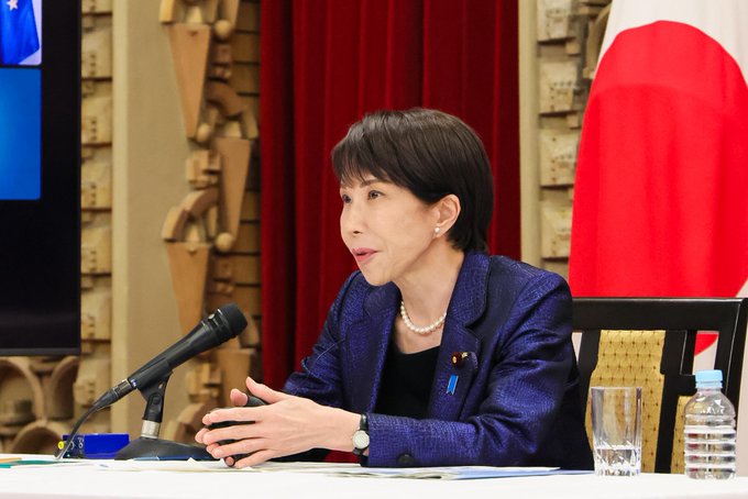 A Japanese official speaks at a press conference, with the Japanese flag prominently displayed. She is likely addressing the financial market downturn discussed in the post.