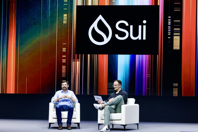 Two men sit on white chairs on stage beneath a large "Sui" logo and water-droplet branding, with one holding a laptop displaying the Sui logo—visually representing the Sui Basecamp event in Singapore where the blockchain was announced to have achieved a record 40 million TPS.