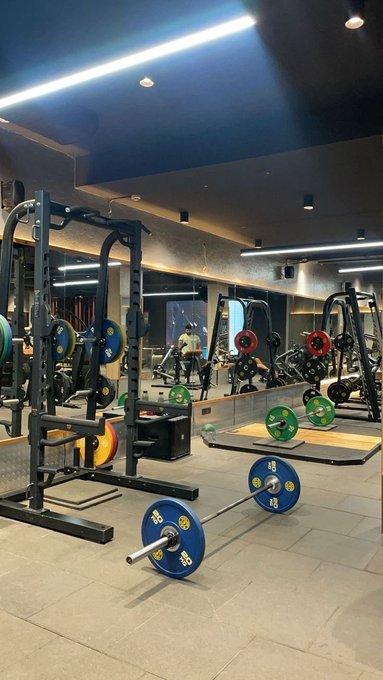 A well-equipped gym featuring squat racks, barbells loaded with blue and yellow weight plates, and lifting platforms on a tiled floor, with mirrors lining the walls reflecting the equipment.