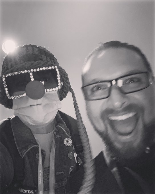 A black-and-white selfie of two people, one wearing a rhinestone-studded cap and a medical mask, the other a bearded man with glasses smiling broadly.