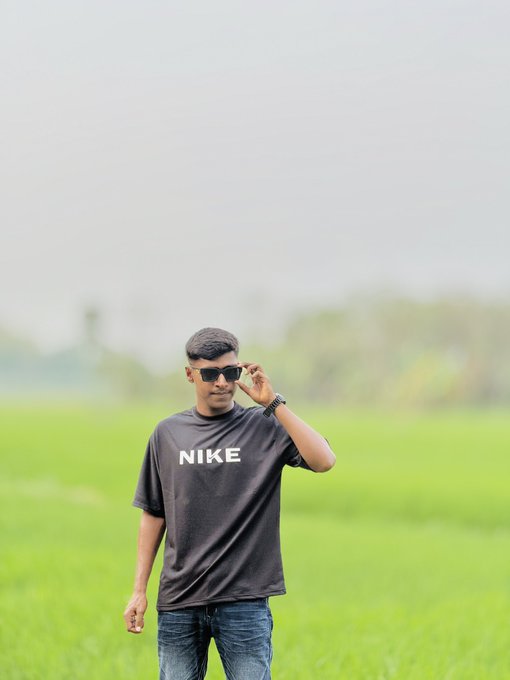 A young man wearing a black Nike t-shirt and sunglasses stands in a lush green field, posing with a peace sign as he cheerfully greets the morning in response to the "gm ☕️" post.