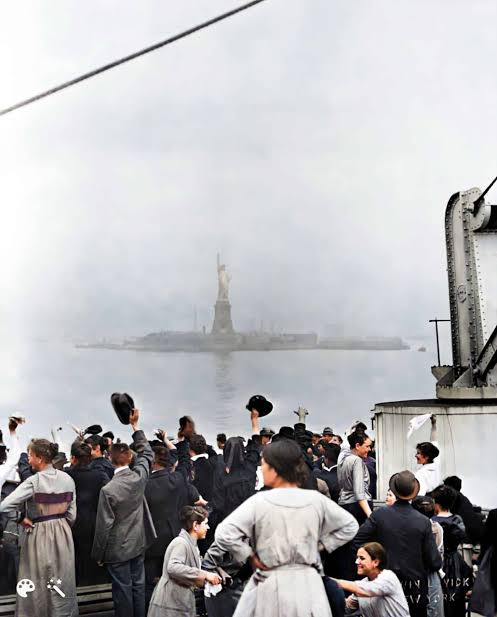 A colorized historical photograph of immigrants crowded on a ship's deck, waving hats toward the Statue of Liberty in the misty background.