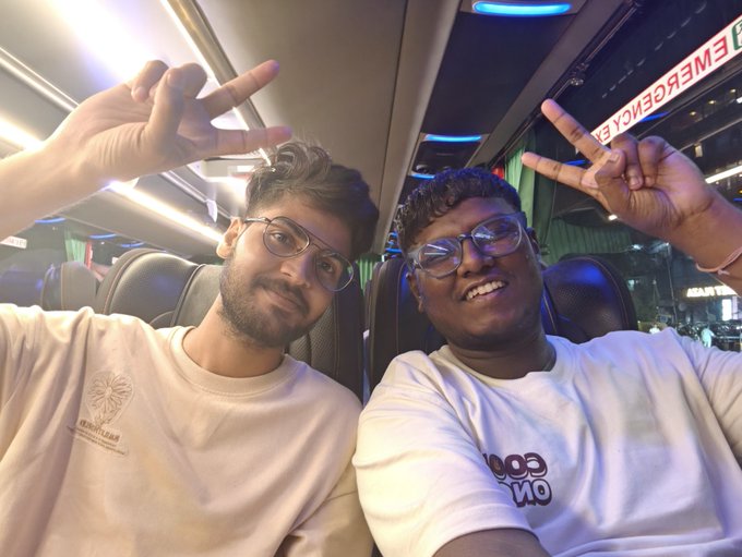A selfie of two young men wearing glasses and white t-shirts, both making peace signs while sitting inside a bus with 'EMERGENCY EXIT' visible.