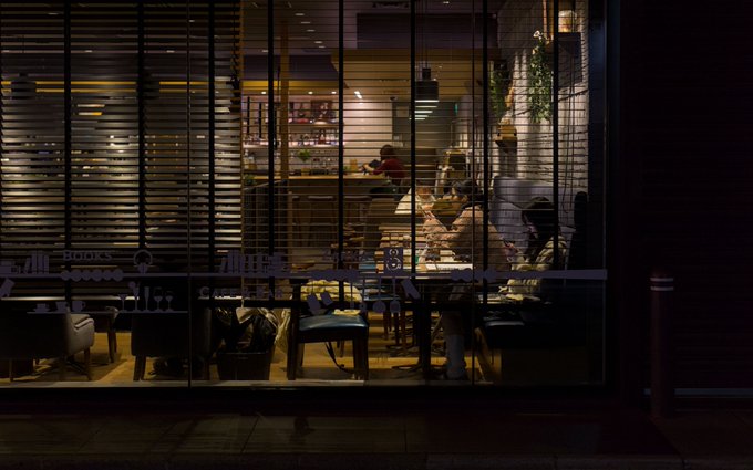 A dimly lit view through slatted blinds reveals the interior of a restaurant or café, with shadowy figures seated at tables and an illuminated bar visible in the background.