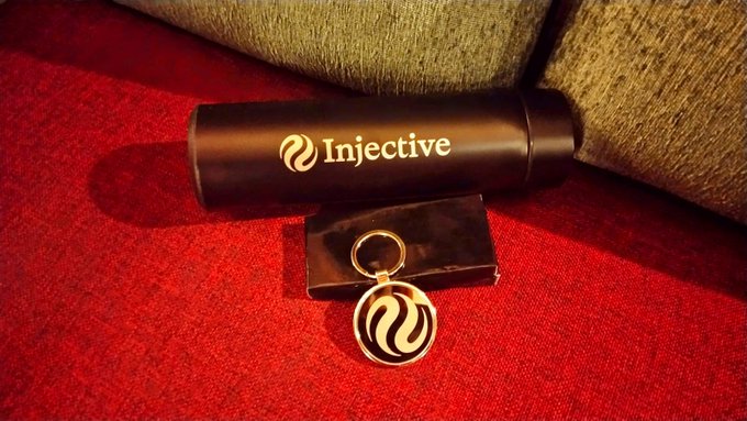 A photograph showing Injective branded merchandise, including a black cylindrical container and a keychain with the Injective logo, displayed on red fabric.
