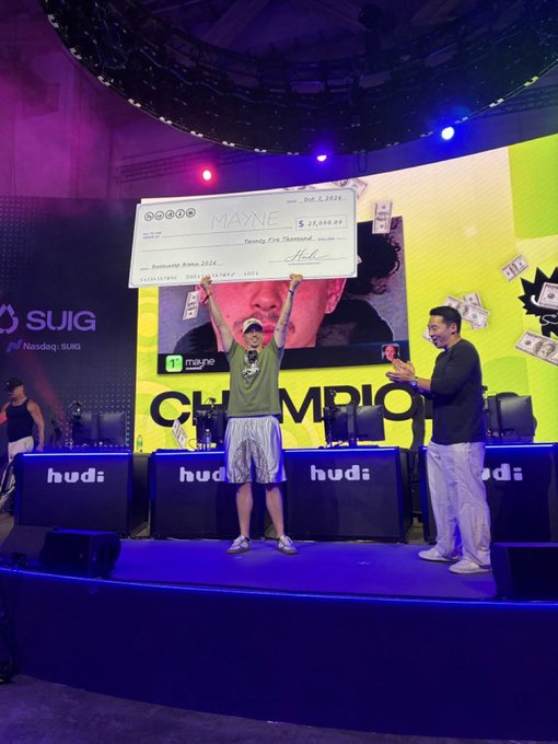 A gaming championship stage where a person holds up a large novelty check made out to 'MAYNE' for $25,000, while another person in a mask applauds, with 'hud:' branding visible on the setup.