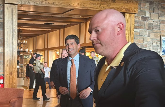 A man in a suit with an orange tie smiles while standing next to a larger man in a dark jacket with a yellow collar, with law enforcement officers visible in the background of what appears to be a lodge or restaurant.
