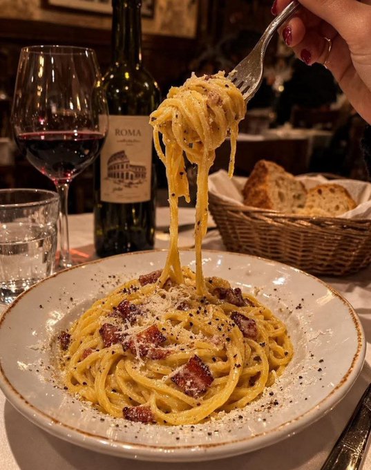 A photo of a plate of carbonara pasta with bacon and cheese being lifted by a fork, accompanied by a glass of red wine, a water glass, a bottle labeled 'ROMA', and a bread basket at a restaurant.