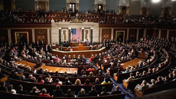 A wide view of the interior of a large, crowded legislative chamber or parliament with an American flag displayed at the front.