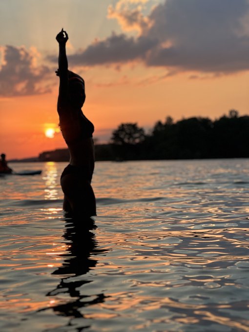 A silhouette of a woman standing waist-deep in calm water at sunset, her arm raised overhead in a yoga pose against an orange sky.