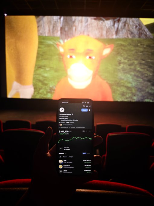 A smartphone in a dark movie theater displaying a crypto portfolio app with a balance of $348,329, with an animated monkey character playing on the large theater screen in the background.