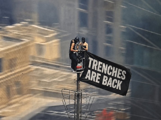 Two people in black ski masks sit atop a pole holding a black banner reading "TRENCHES ARE BACK" in bold white letters, overlooking a city street—evoking a return to public spectacle and protest activity after a long lull.