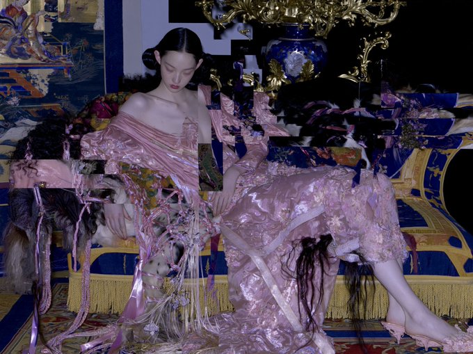 A stylized fashion editorial image shows a model in an ornate pink dress with intricate embroidery and fringe details, seated on a luxurious gold and blue baroque-style chaise lounge.