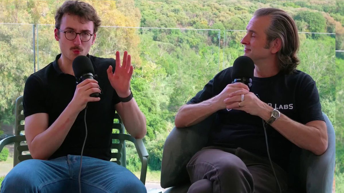 The image shows two men sitting in chairs, each holding a microphone. They appear to be engaged in conversation or discussing something related to technology. The background features a window overlooking a forest, adding an element of nature to the scene. The caption at the bottom of the image reads "Zcash and the Threat of Quantum Computers New breakthroughs are coming out at an accelerating pace". This suggests that the men might be talking about recent advancements in technology or discussing the implications of these developments on society and security.