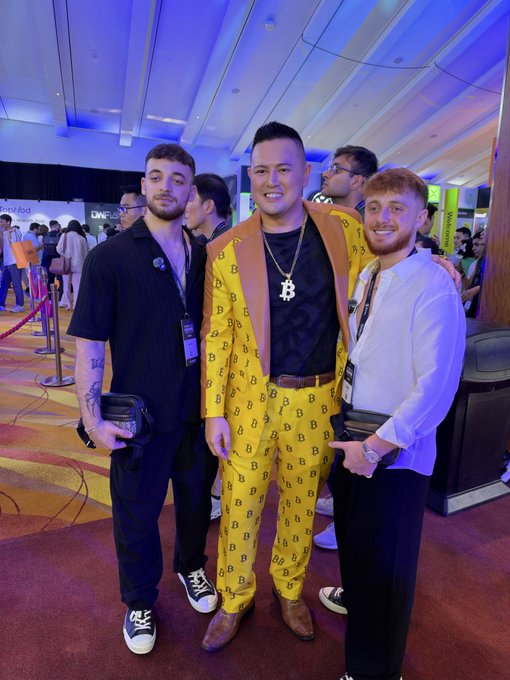 Three men posing together at what appears to be a crypto conference; the man in the center wears a yellow Bitcoin-patterned suit and a Bitcoin necklace, with banners for Polygon and other projects in the background.