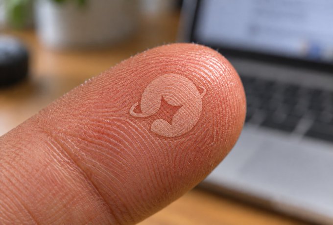 Close-up macro photograph of a human fingertip displaying a small, raised, light-colored tattoo of a logo resembling a stylized 'C' with a star inside, set against a blurred laptop background.