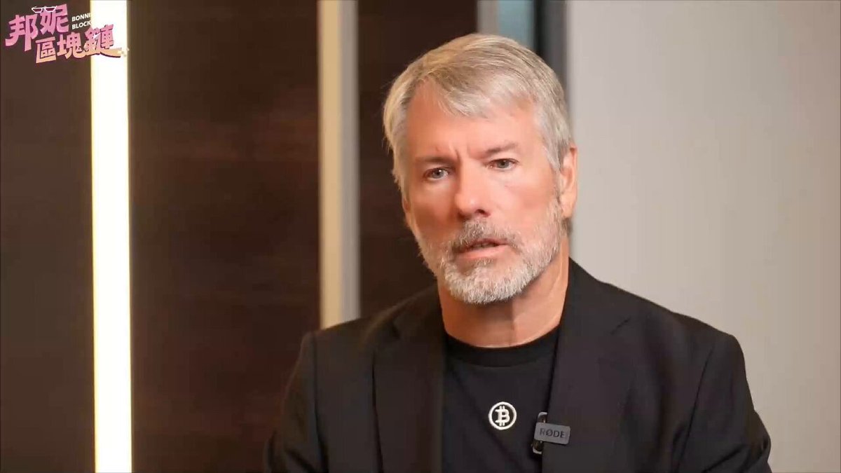 The image features an older man wearing a black suit jacket with a white beard sitting at a desk or table. He appears to be looking directly into the camera while seated. The background of the image shows a wall, which could potentially be part of a room in a building. There is also text visible on the image that reads "Strategy Selling 32 Bitcoins Sparks Widespread Online Discussion Michael Saylor's previous video has already explained this logic: Saylor'S original words were 'Never become a net seller of Bitcoin—selling 1 coin while simultaneously buying 10 to 20, the Strategy is still net'. Focus on what is in the image and how it relates to the post."