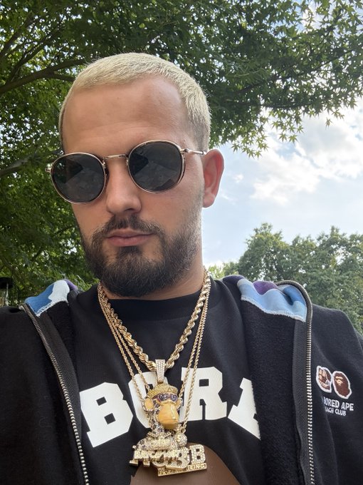 A selfie of a man with short blonde hair, a beard, and round sunglasses, wearing a black BAPE hoodie and multiple gold chains.