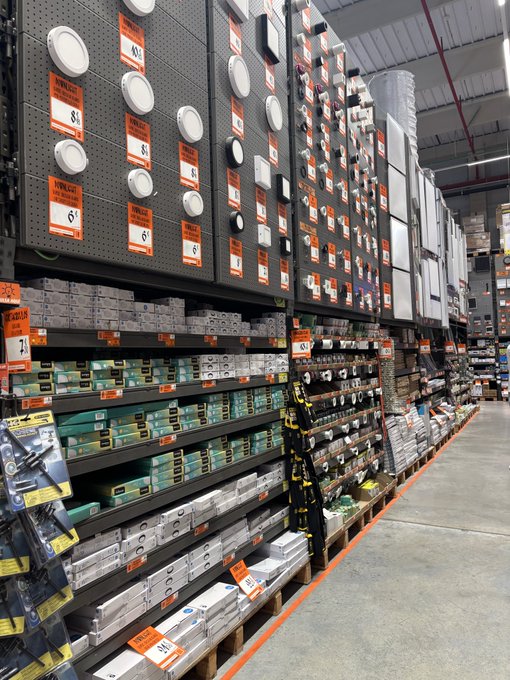 A worker stands exhausted in a long aisle of a Home Depot warehouse, surrounded by towering shelves of light fixtures and hardware priced at $6, $7, $8, and $34, visually embodying the weariness of spending every Saturday stocking these endless rows of inventory.