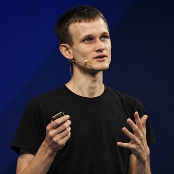 A man with short hair and a goatee, wearing a black t-shirt and a headset microphone, speaks while gesturing with his hands and holding a small yellow device, appearing to deliver a presentation against a blue backdrop. The image aligns with the post's context, as the speaker resembles Vitalik Buterin, the Ethereum co-founder, sharing his views on education reform.