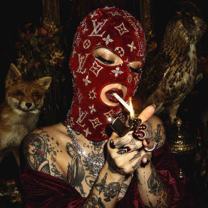 A woman wearing an ornate red Louis Vuitton-patterned mask with rhinestones, lighting a cigarette while a fox and owl sit in the background amid dark, baroque surroundings.
