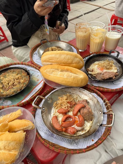 A table filled with Vietnamese cuisine, featuring banh mi sandwiches and bowls containing eggs, sausages, meat, and noodles, accompanied by iced coffee drinks.