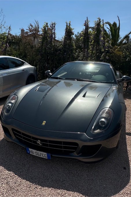 A grey Ferrari sports car with a French license plate (HB-367-CC) parked on a gravel driveway with greenery in the background.