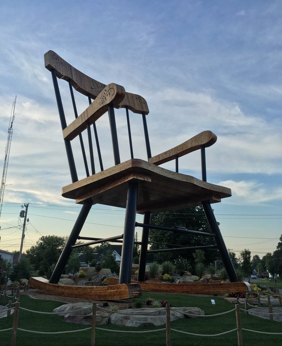 A massive wooden rocking chair sculpture displayed outdoors as a roadside attraction, towering over the landscaped area beneath a cloudy blue sky.