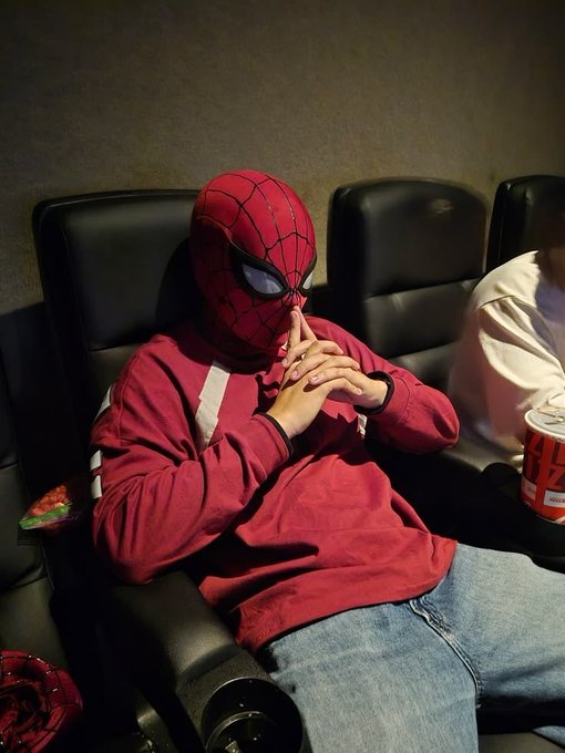 A person wearing a Spider-Man mask and a red sweatshirt sitting in a black leather chair with their hands clasped near their face, with a bowl of snacks and a drink cup visible beside them.