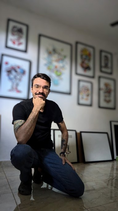 A man with glasses and a mustache kneels thoughtfully on a tiled floor in front of a wall displaying a gallery of framed artwork, projecting a creative, OG artist-at-work vibe. The casual pose and studio backdrop pair well with the "GM to the OGs" greeting, evoking an artistic salutation to longtime supporters.