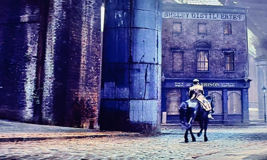 A lone figure in a long coat and hat rides a dark horse down a dimly lit cobblestone street, passing a brick building with painted signage reading "SHELLEY DISTILLERIES" and "HARRISON WINE & SPIRITS."
