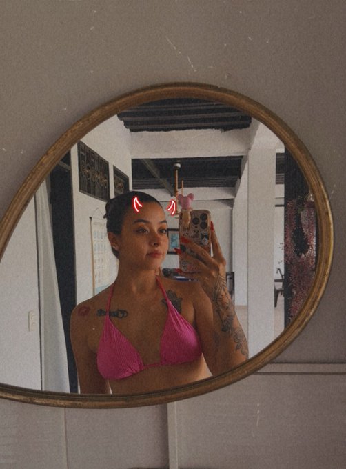 A mirror selfie of a woman with her hair in a bun wearing a pink bikini top, featuring a devil horn filter and showing a heart tattoo on her chest.