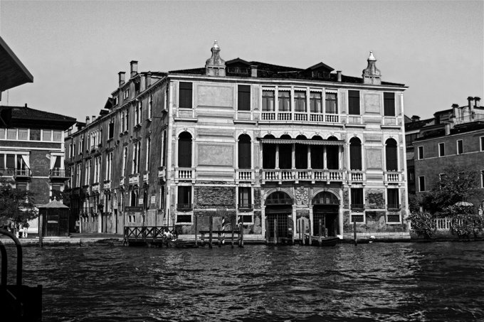 A black and white photograph showcasing an elegant Venetian palazzo rising directly from the Grand Canal, its classical façade featuring arched windows, balconies, and decorative rooftop finials that embody timeless waterfront sophistication. The architectural beauty pairs perfectly with the post's "Elegance by the Water" theme, evoking the refined charm reflected in the $XTZ token's presentation.