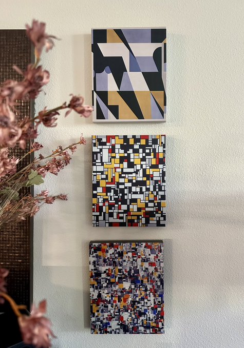 Three geometric abstract paintings in black, white, gold, red, blue, and gray hang vertically on a white wall next to pink flowers.
