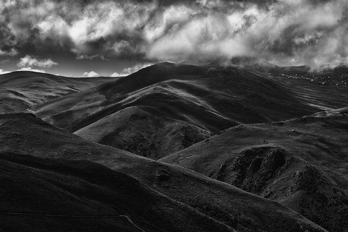 A dramatic black and white photograph of rolling mountain hills under a sky filled with heavy, textured clouds.