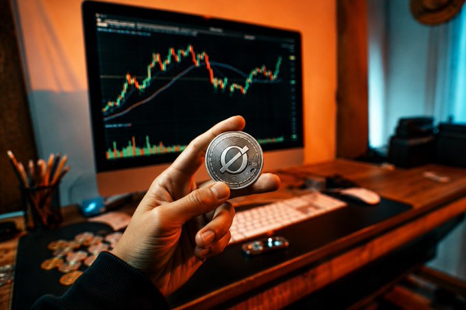 A hand holding a physical Stellar (XLM) cryptocurrency coin in front of a computer monitor displaying a candlestick stock or crypto price chart.