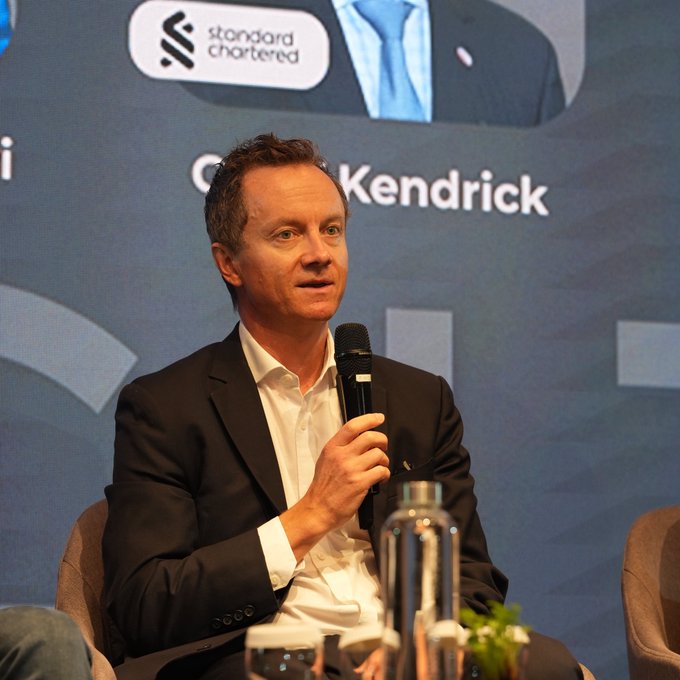 A man identified as "Kendrick" speaks into a microphone at a Standard Chartered event, with the "standard chartered" logo visible in the background. The image relates to the accompanying text, showing Geoff Kendrick, the Standard Chartered analyst, delivering his prediction that the entire foreign exchange market could shift to stablecoins by 2030.