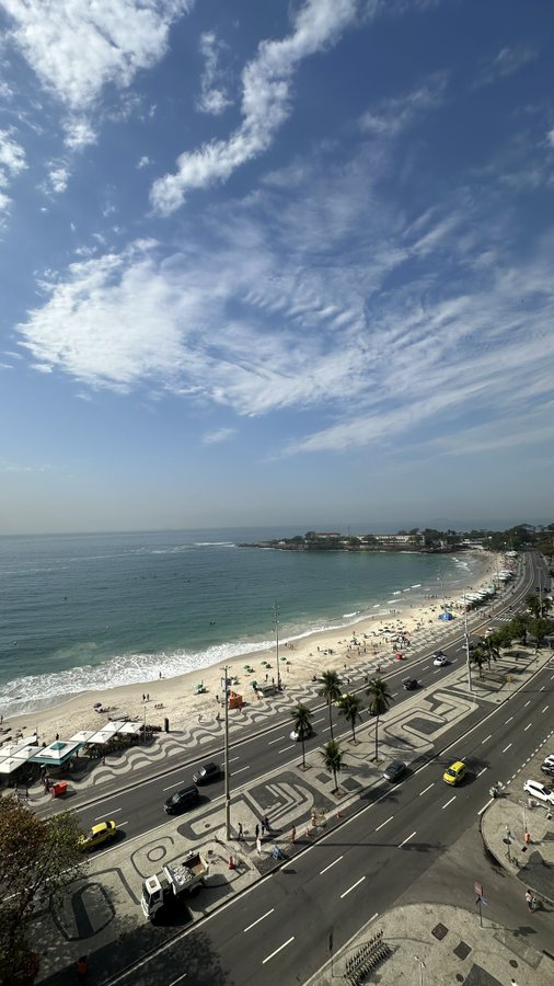 An aerial, wide-angle view overlooks a coastal city, showing a sandy beach beside a road with Portuguese-style mosaic pavement under a vast, cloudy sky.
