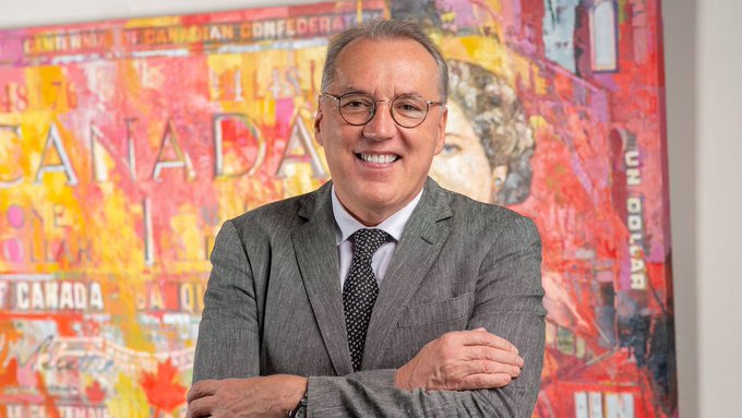 The image features a man wearing glasses and a suit, standing next to a painting of a woman's face with a Canadian sign behind him. The man appears to be smiling for the camera, possibly posing for a portrait or an advertisement. The painting is located in front of a wall that has a "CANADA" sign on it, indicating its connection to Canada. The overall scene suggests that the man might be involved in some business related to the Canadian market or industry, as he is dressed professionally and standing next to a piece of art with a Canadian theme.