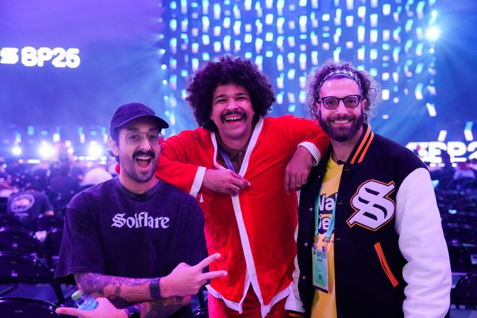 The image shows three men posing together, with one man wearing a red jacket and another wearing a black jacket. They are standing next to each other against a purple background, which appears to be part of a stage or concert venue setup. The third person is also present in the scene but only partially visible. In the foreground, there's a caption that reads "Reminder: Solana is you You want to stand out and feel supported by Solana? Show up IRL and give the ecosystem a real person to root for". This caption suggests that the image might be related to an event or concert featuring the Solana cryptocurrency.