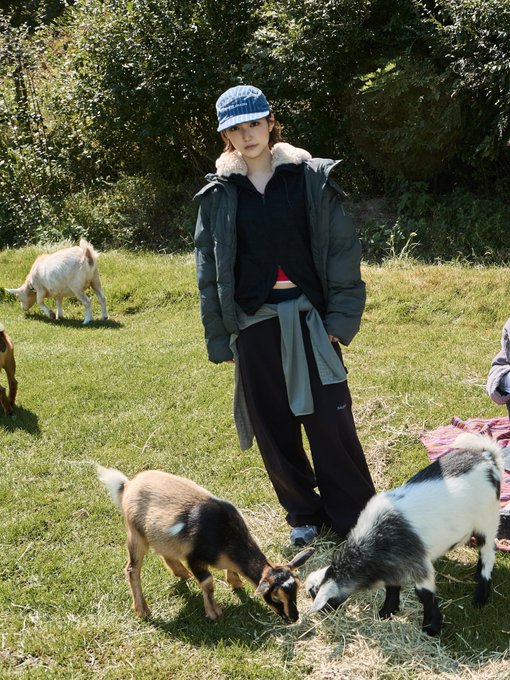 Full-body shot of a young woman wearing a blue bucket hat, a black puffer vest over a black hoodie, and black pants, standing in a grassy field surrounded by several goats.
