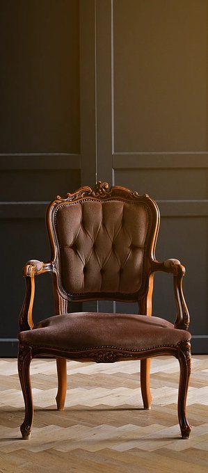 An ornate antique wooden armchair with brown tufted upholstery sits on a chevron-patterned parquet floor against a dark paneled wall.