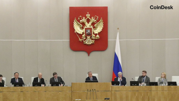 The image shows officials seated at a dais beneath the Russian Federation's state emblem and flag, with "CoinDesk" visible in the corner. This visual context relates to the accompanying post about Russia's State Duma advancing revised cryptocurrency oversight legislation that relaxes wallet disclosure requirements, caps retail crypto investments at 300,000 rubles per year, and imposes a 48-hour delay on large foreign transfers.