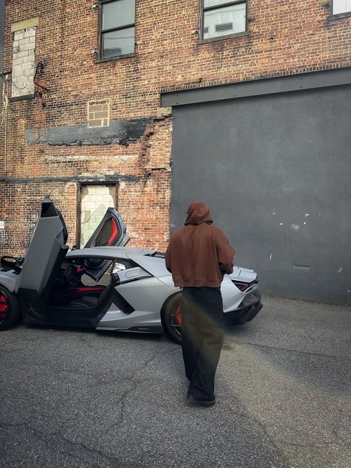 A person in a brown hooded jacket stands beside a silver Lamborghini with its scissor door open, parked against a weathered brick building.