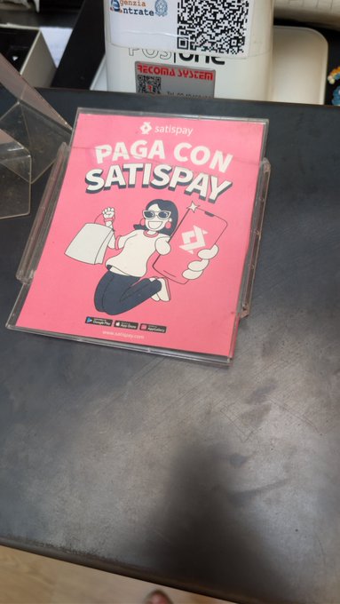 A pink placard with the text 'PAGA CON SATISPAY' and a cartoon illustration sits on a counter in front of QR codes labeled 'intrate' and 'REKORA SÜZÜM'.