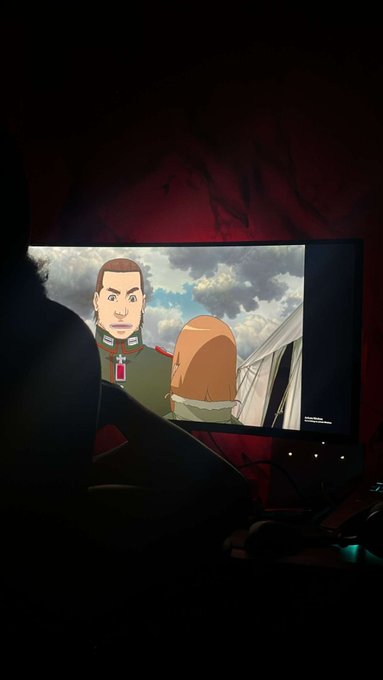 A person watches a screen displaying an anime character with the insignia 'RED' on their collar, visible against a dark, dimly lit background.