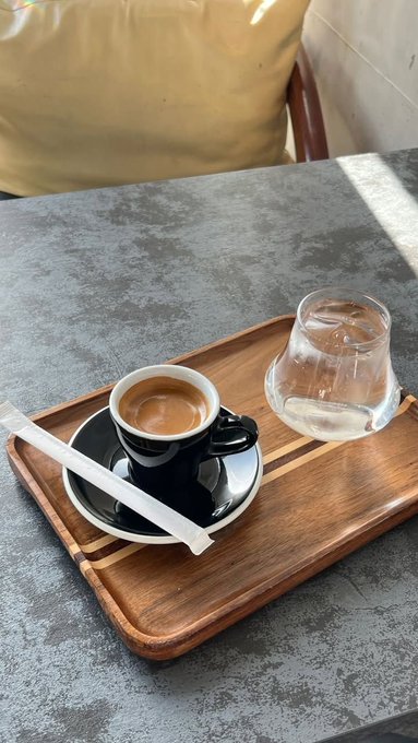 A small black espresso cup with a straw and an empty glass of water served on a wooden tray atop a grey stone table.
